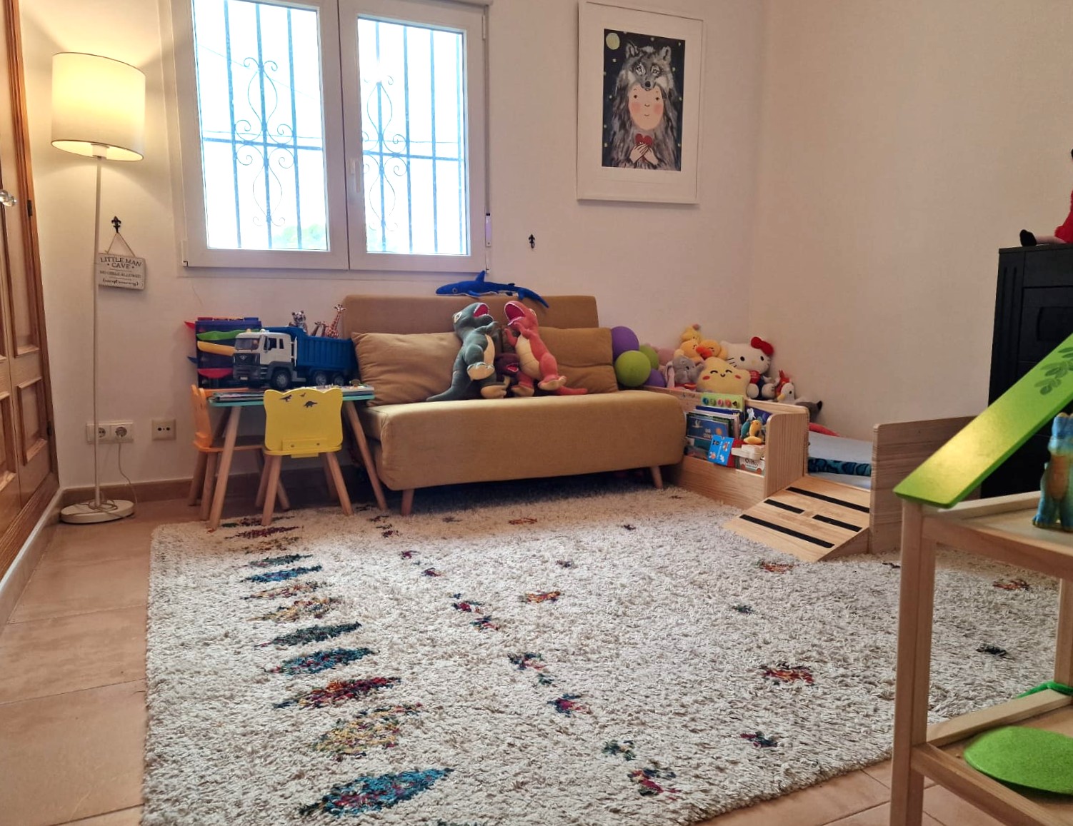 Espace de jeux pour enfants avec un tapis texturé, un canapé jaune et une table d'activités. La pièce dispose de lumière naturelle et d'une fenêtre avec grilles de sécurité.