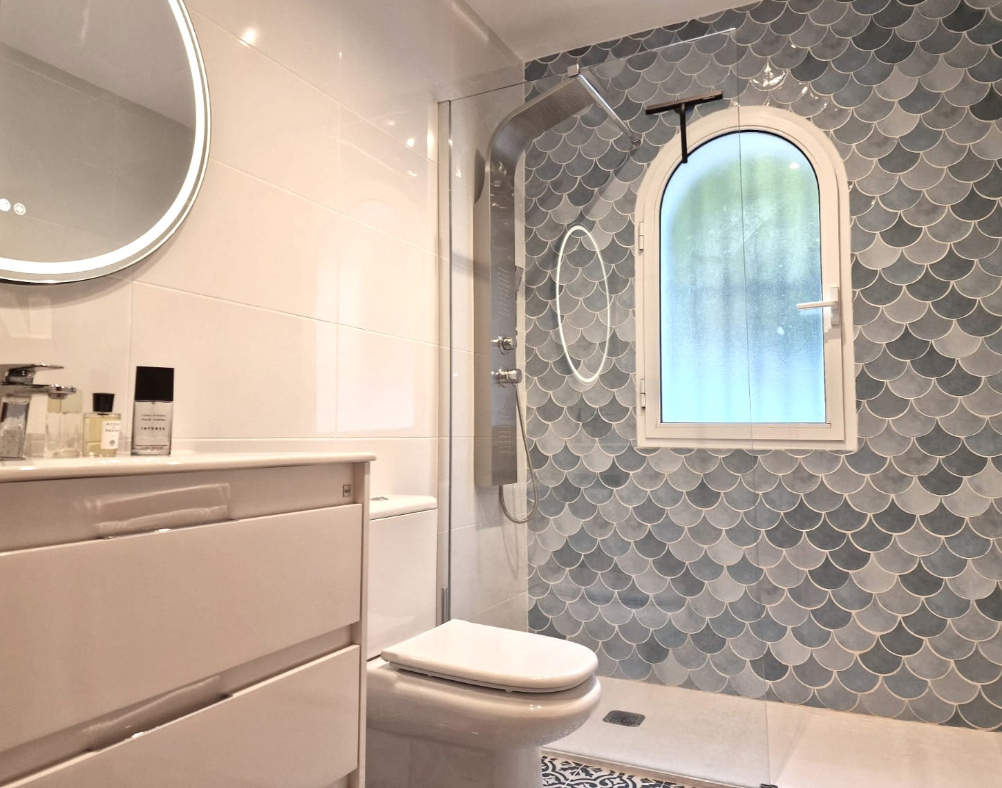 Salle de bains moderne avec douche à l'italienne, carrelage en écailles bleues, fenêtre en arche dépolie, colonne de douche en inox et miroir circulaire rétroéclairé.