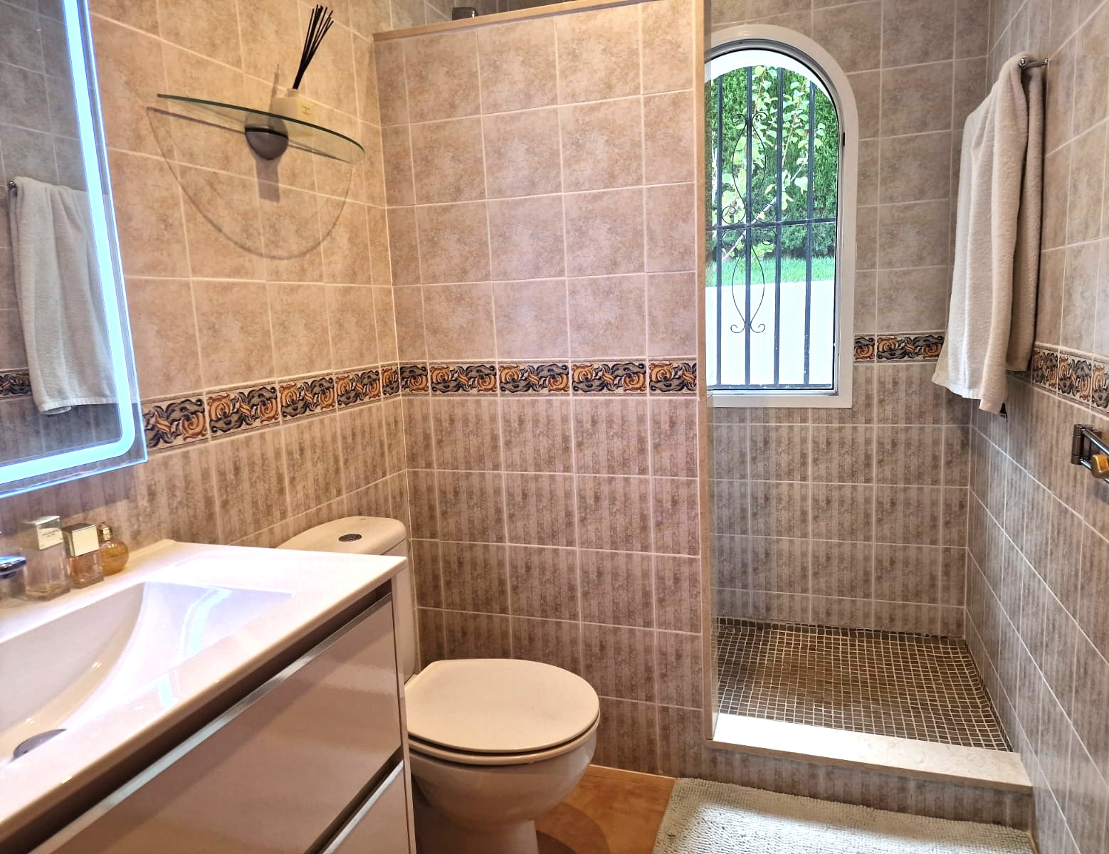 Salle d'eau moderne avec douche à l'italienne, carrelage beige orné d'une frise décorative et fenêtre en plein cintre offrant une luminosité naturelle.