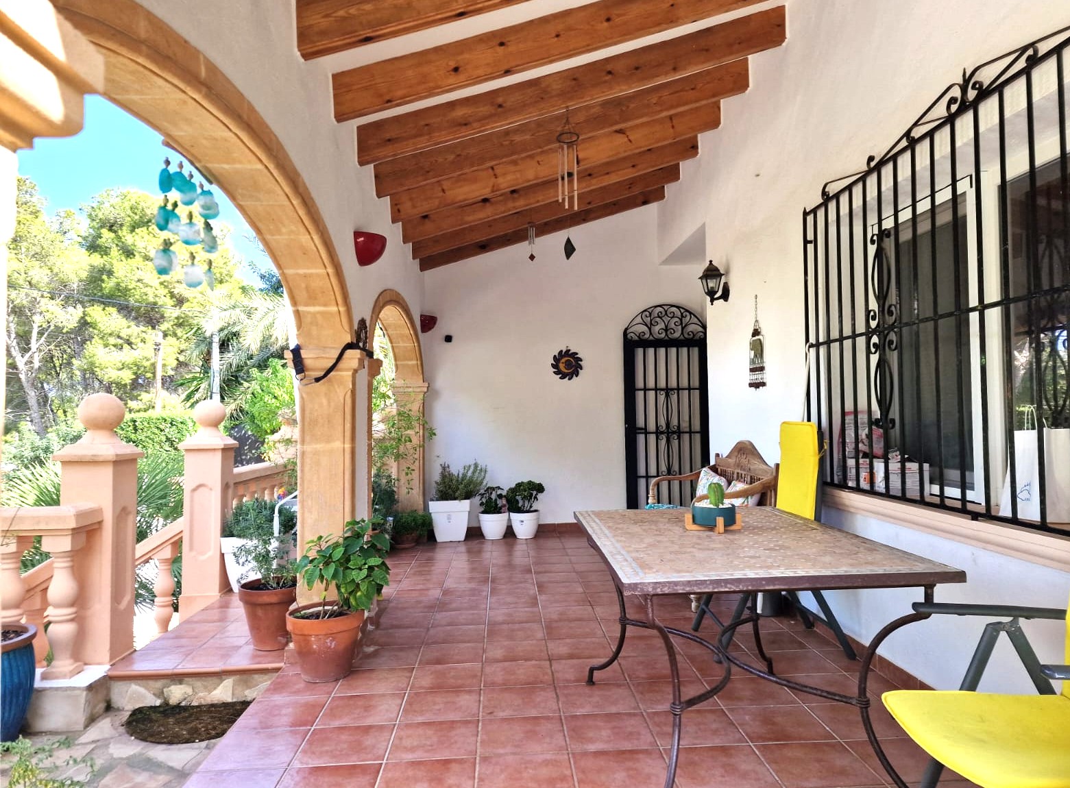 Terrasse couverte avec arches en pierre de Tosca, poutres en bois apparentes et sol en terre cuite. Espace extérieur avec coin repas et vue sur la végétation.