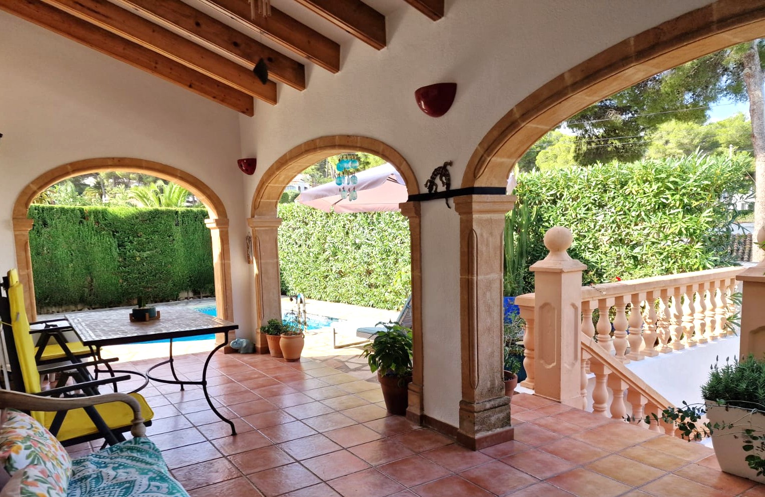 Terrasse couverte avec arches en pierre de Tosca, poutres en bois apparentes et sol en terre cuite, offrant une vue sur la piscine et le jardin.