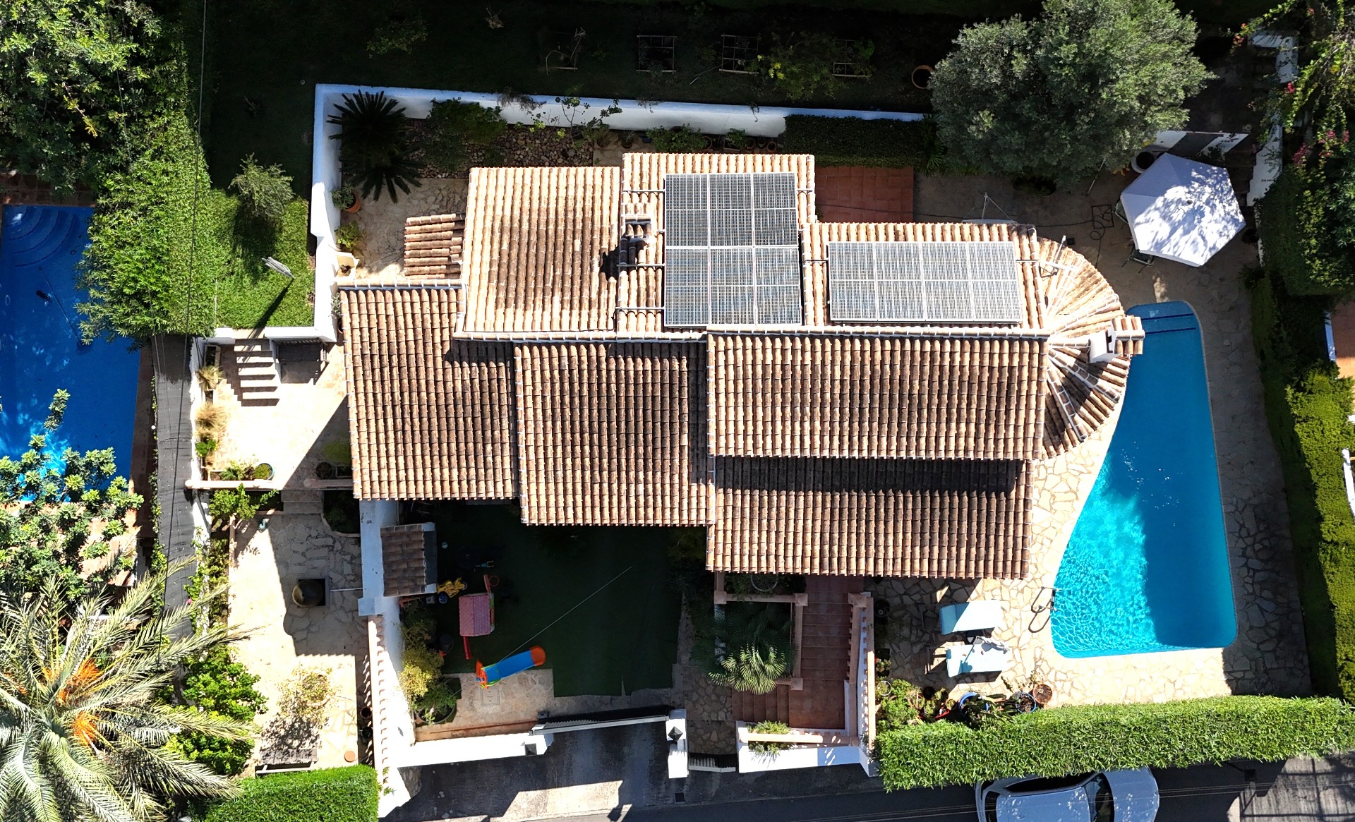 Vue aérienne de la villa présentant le toit en tuiles avec panneaux solaires, la piscine privée et un jardin méditerranéen avec espace de jeux pour enfants.