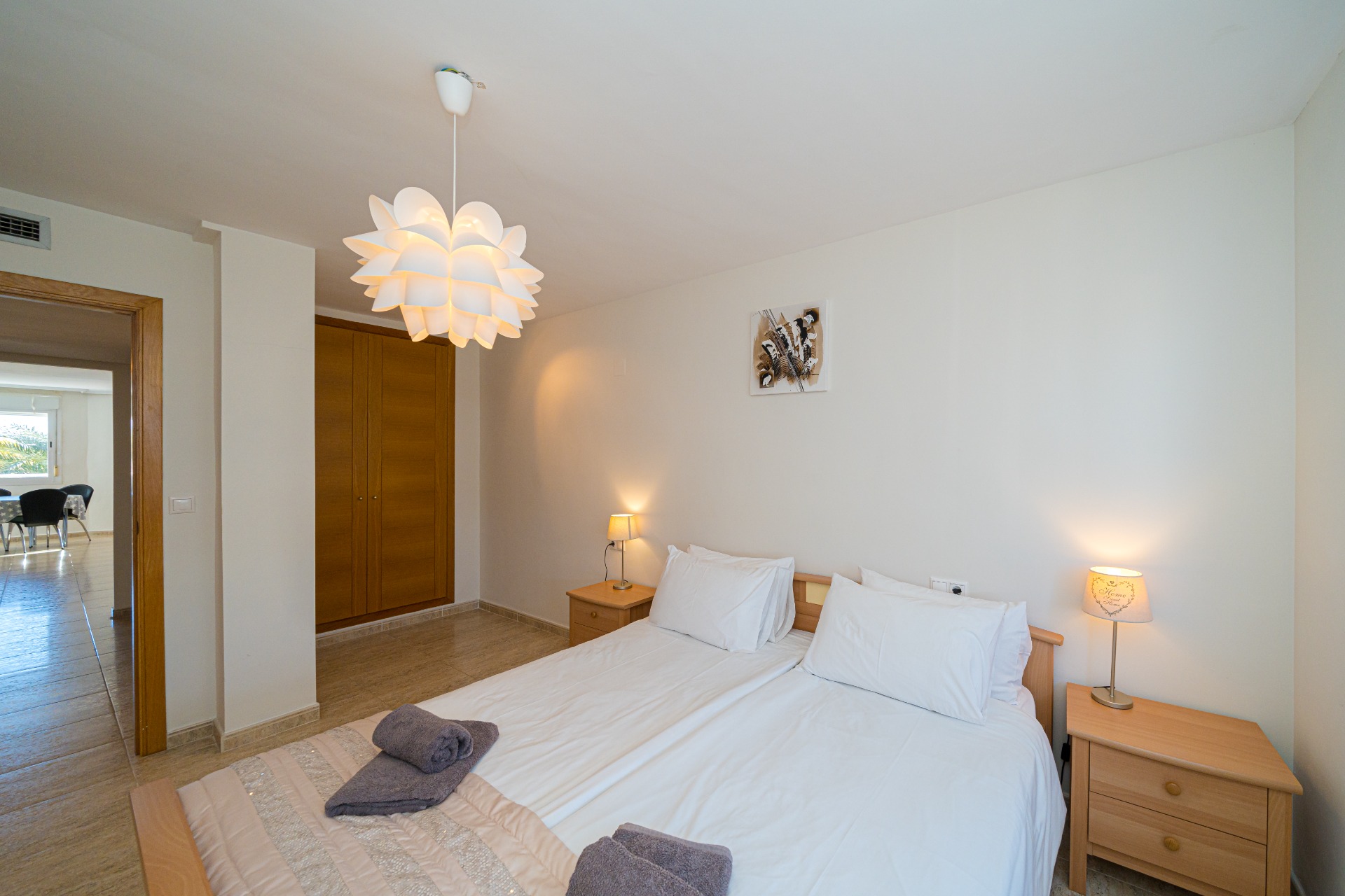 Chambre avec lits jumeaux, armoires encastrées en bois et luminaire suspendu en forme de fleur. La porte ouverte offre une vue sur la salle à manger lumineuse.