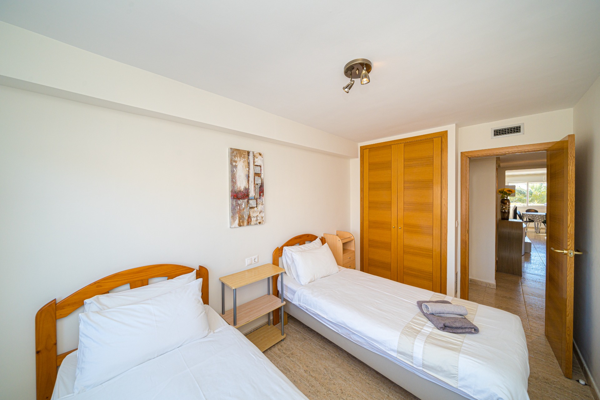 Chambre lumineuse avec deux lits jumeaux, têtes de lit en bois, armoire encastrée en chêne et sol en grès. Une porte ouverte mène vers l'espace de vie.