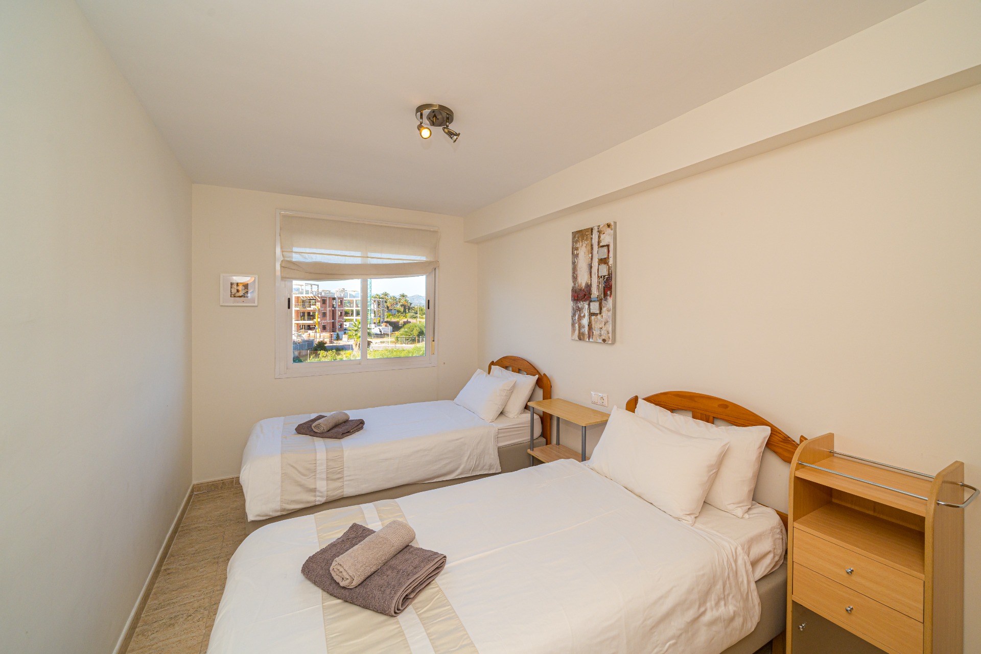 Chambre lumineuse avec deux lits simples, têtes de lit en bois, sol en carrelage et une fenêtre offrant une lumière naturelle et une vue sur les environs.