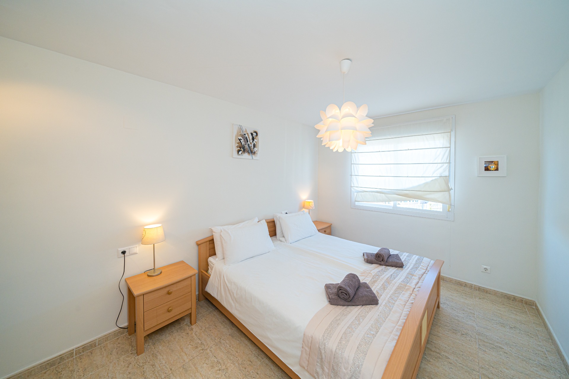 Chambre lumineuse avec mobilier en bois clair, lit double et tables de chevet assorties. La lumière naturelle pénètre par la fenêtre à store blanc sur un sol en grès.