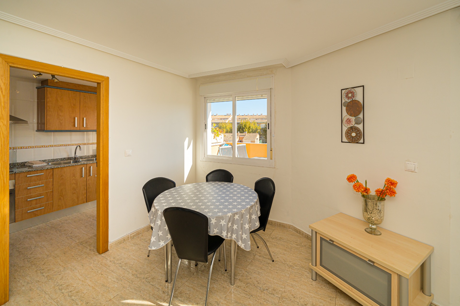 Coin repas lumineux avec table ronde et carrelage au sol, attenant à une cuisine fonctionnelle en bois. La fenêtre offre une vue sur les environs de Javea.