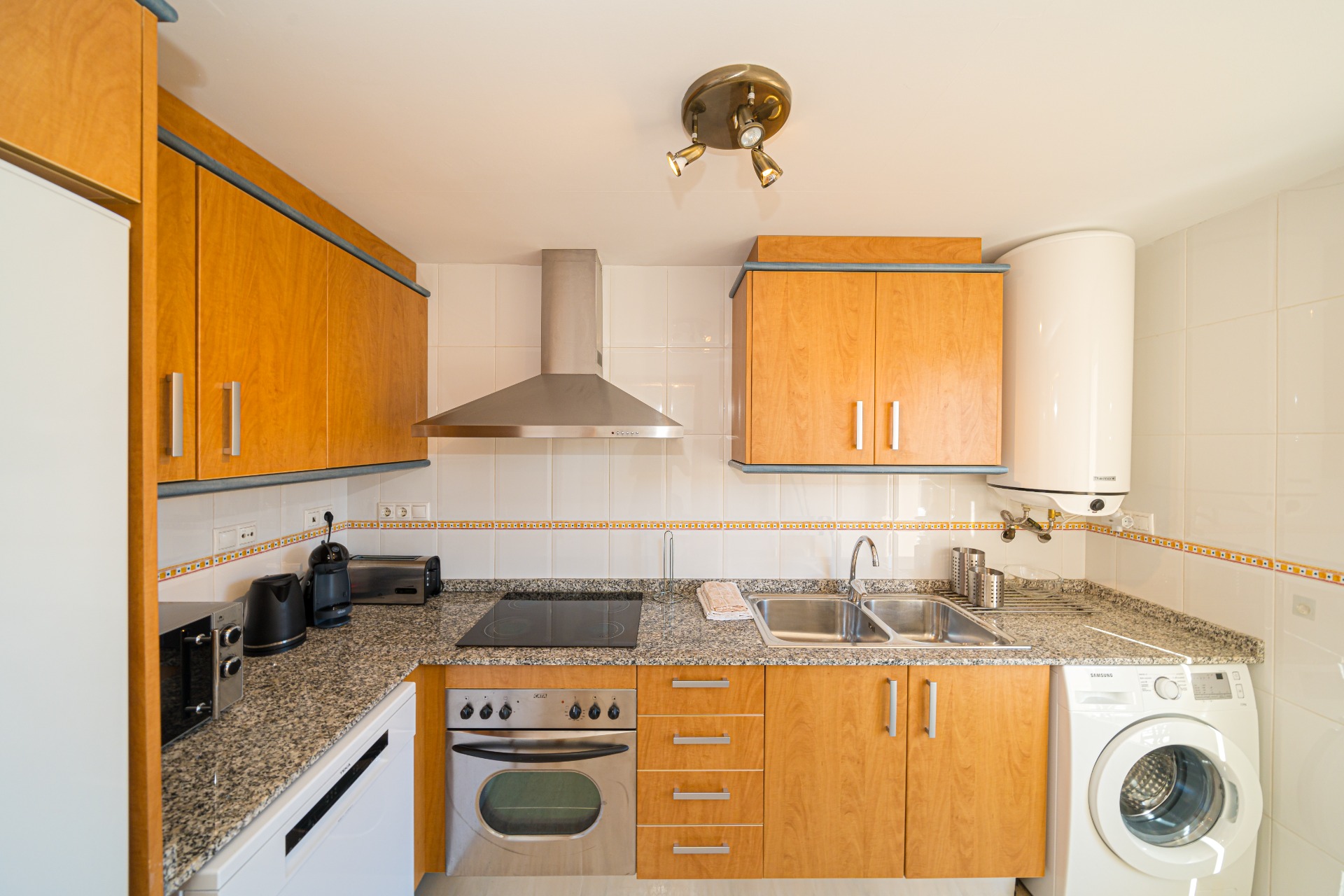 Cuisine équipée avec meubles en bois, plan de travail en granit et électroménagers intégrés, incluant un four, un lave-linge et une hotte en inox.