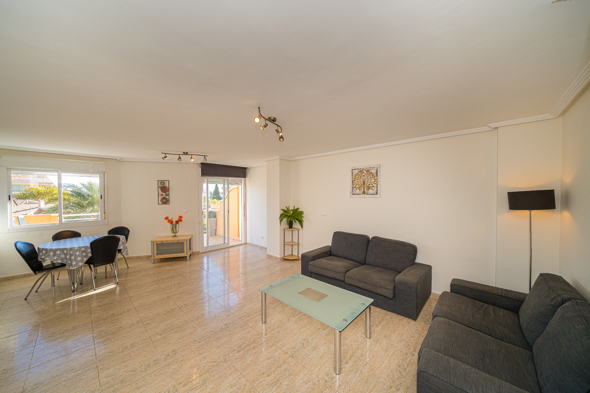 Espace de vie ouvert comprenant un salon avec canapés gris, un coin repas et un sol en grès cérame. Les baies vitrées offrent un accès direct à la terrasse extérieure.