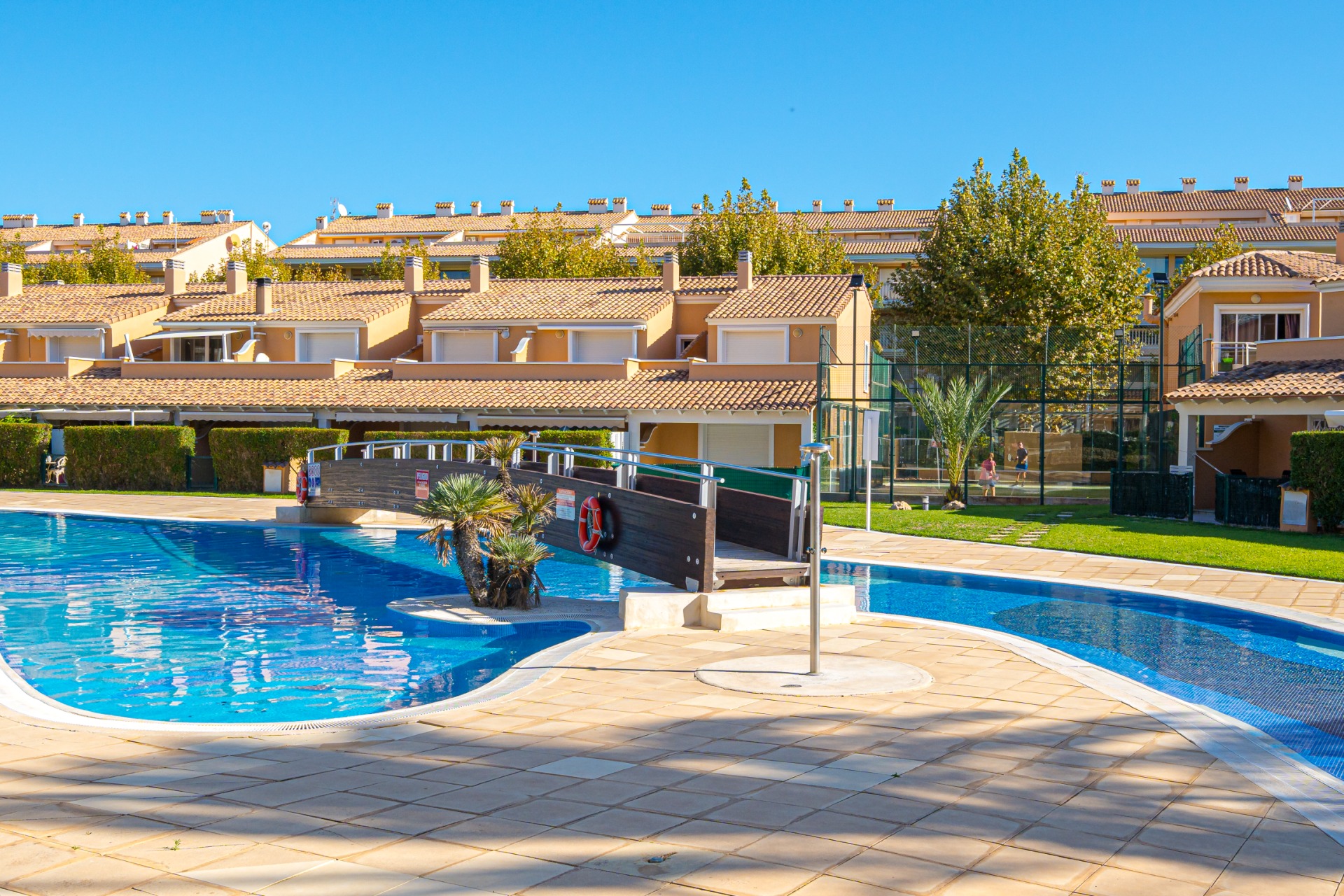 Grande piscine communautaire avec pont en bois, entourée d'une terrasse ensoleillée et d'un court de padel en arrière-plan du complexe.