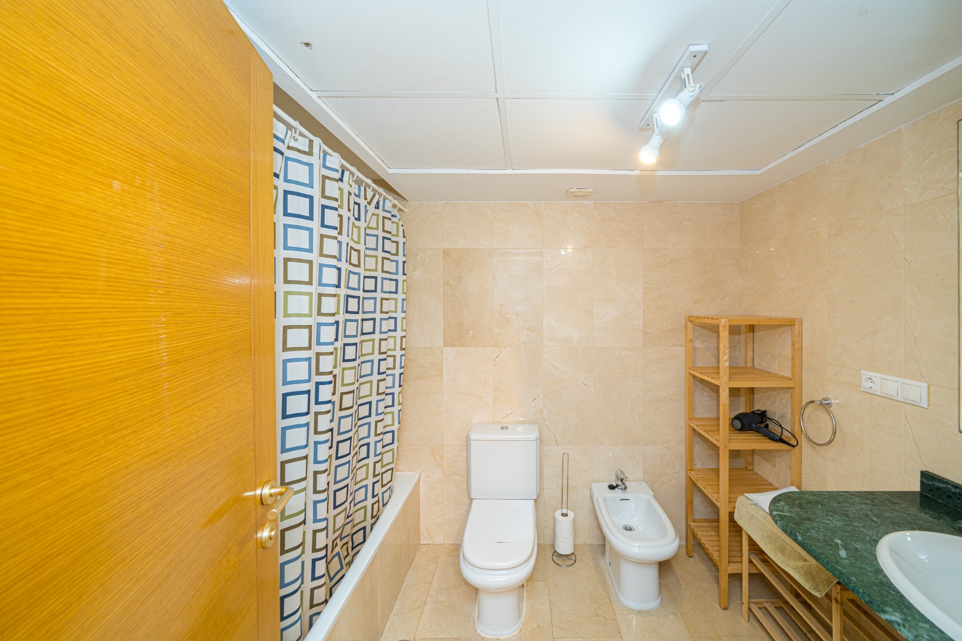 Salle de bain carrelée en marbre beige comprenant une baignoire, un bidet et un plan de toilette en marbre vert avec des étagères en bois naturel.