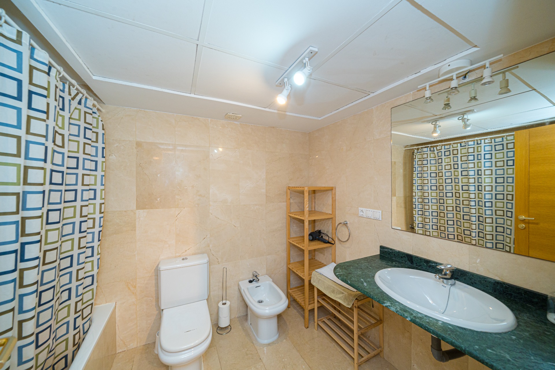 Salle de bain moderne avec carrelage en marbre crème, plan de toilette en marbre vert et bidet. Équipée d'étagères en bois et d'un éclairage sur rail au plafond.