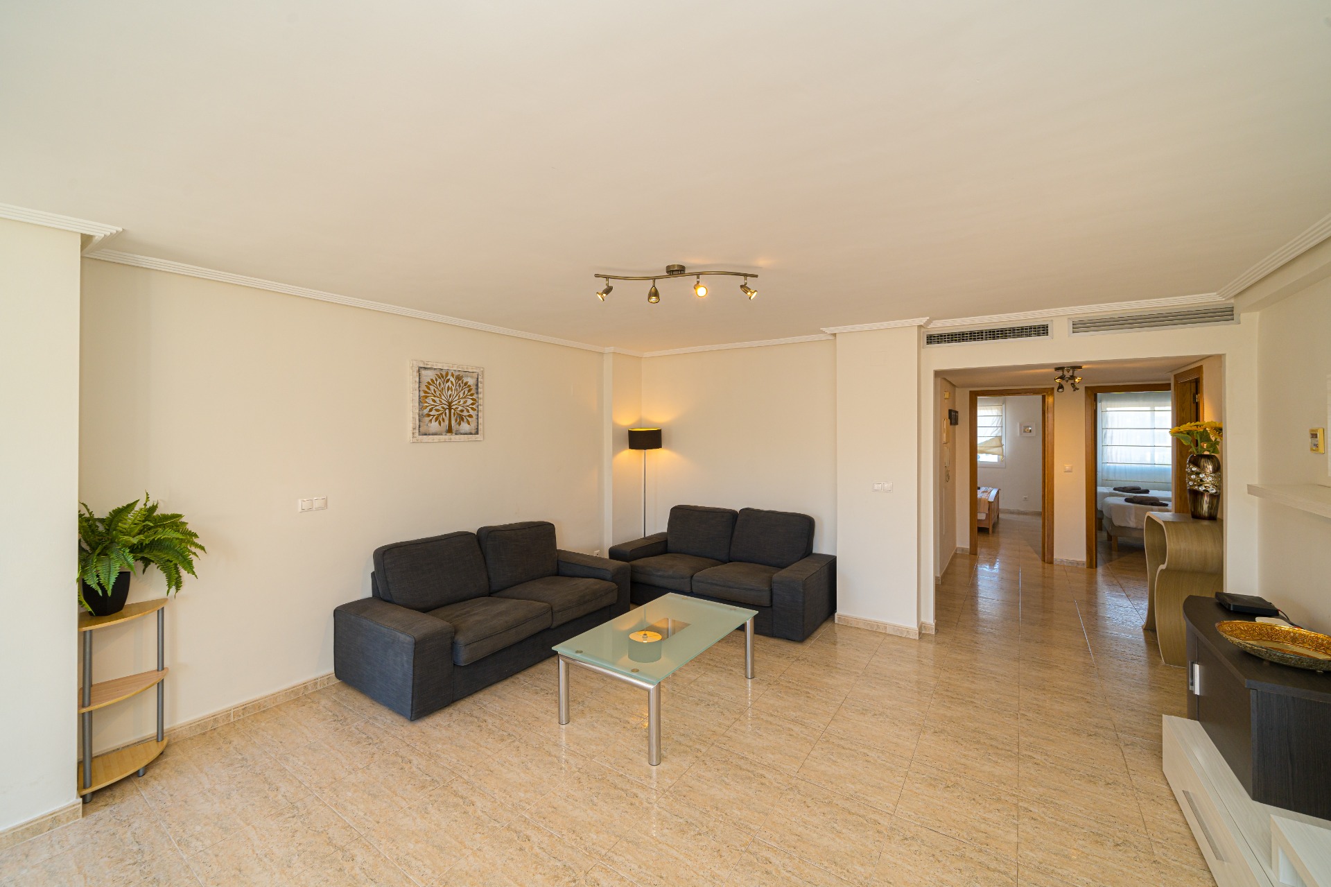 Salon spacieux avec canapés gris foncé, table basse en verre et sol en carrelage poli. La disposition ouverte mène vers le couloir desservant les chambres.
