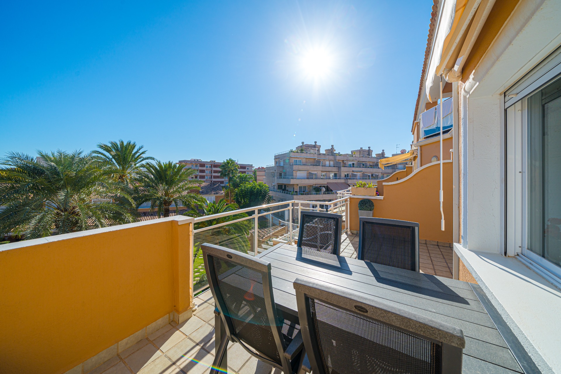 Terrasse privée ensoleillée avec coin repas extérieur gris foncé, sol carrelé et vue sur les palmiers et les résidences environnantes.