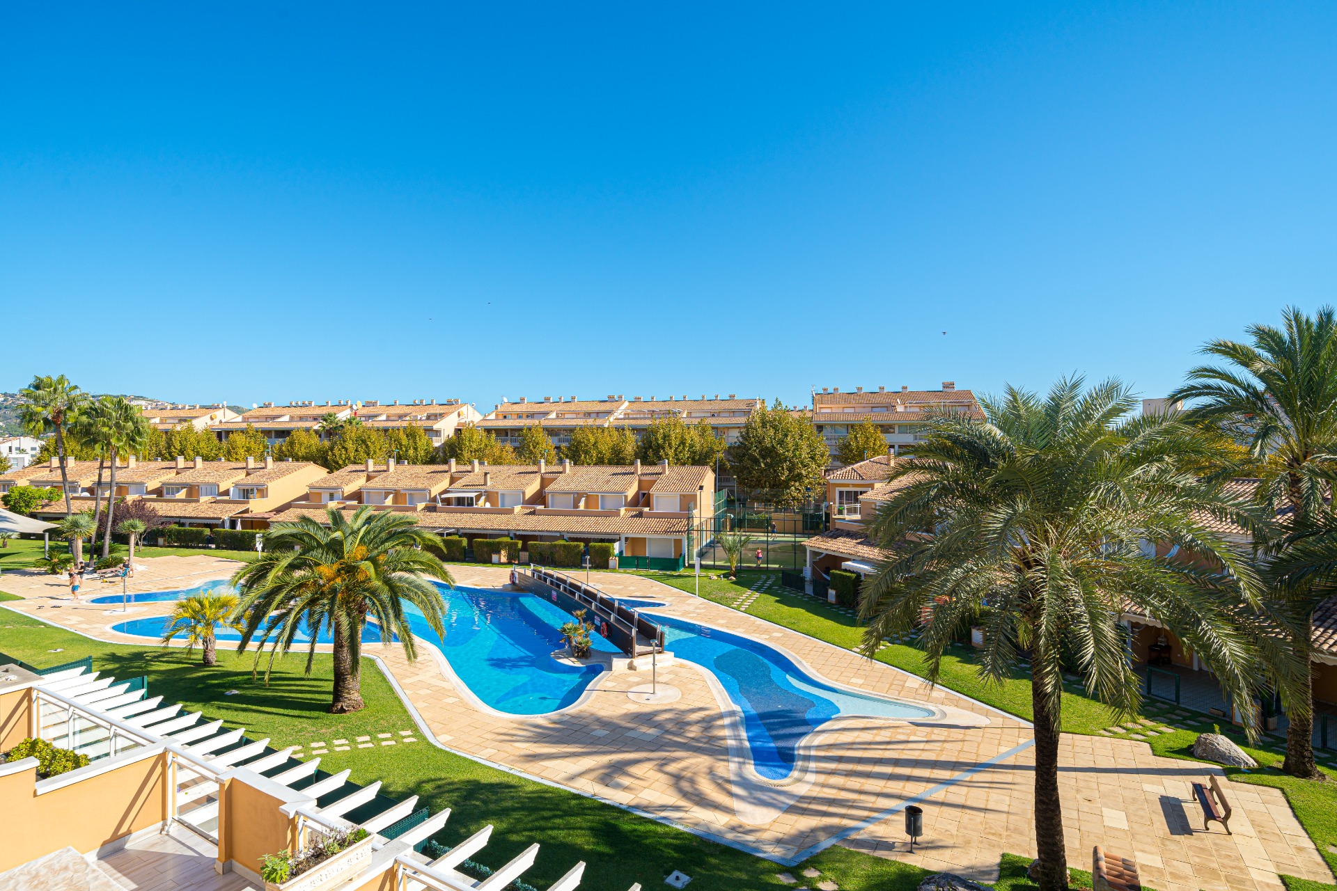 Vue sur la grande piscine communautaire en forme de lagon avec pont central, entourée de palmiers et de jardins paysagers au sein du complexe Moreras del Saladar.