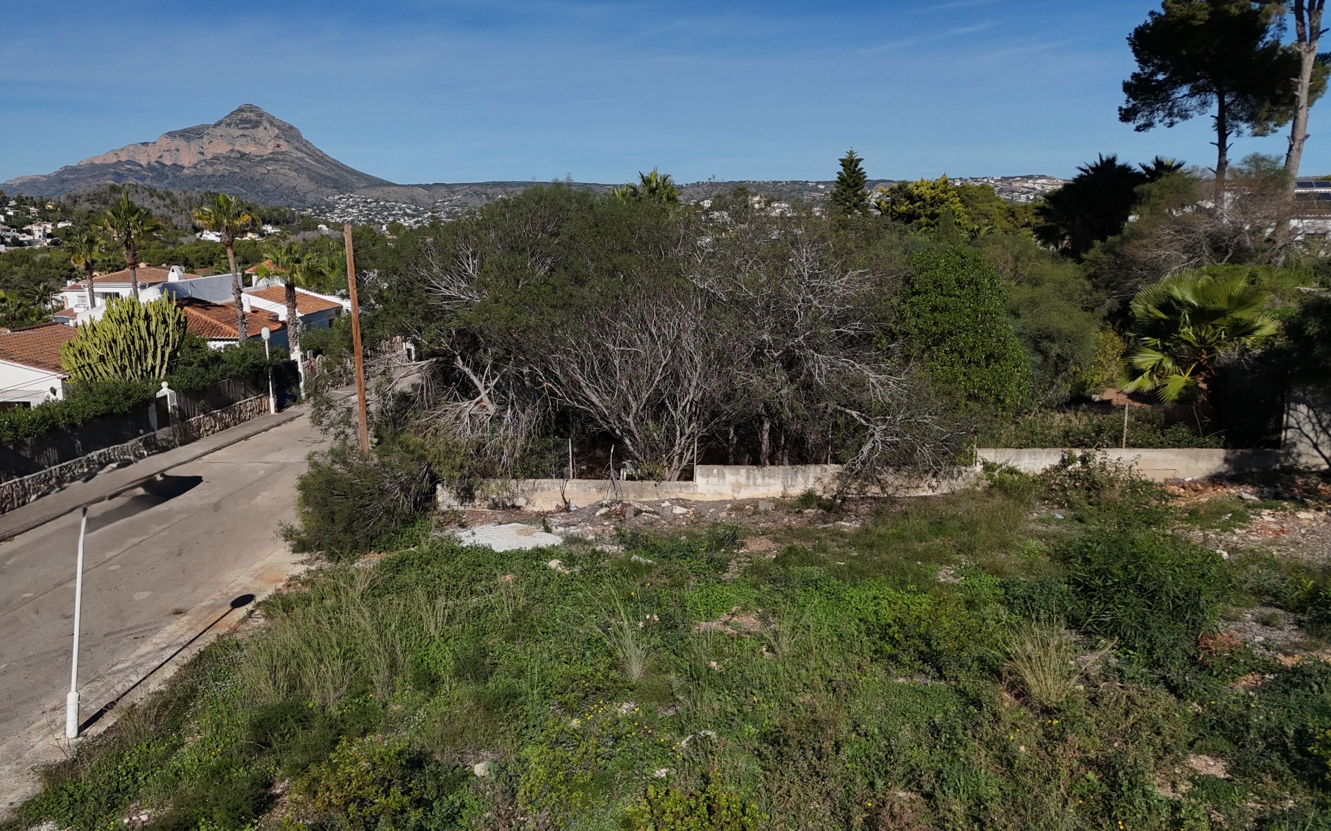 Vue aérienne du terrain plat de 889 m² à Pinosol, montrant la rue résidentielle calme, les murs de clôture et une vue dégagée sur la montagne Montgó.