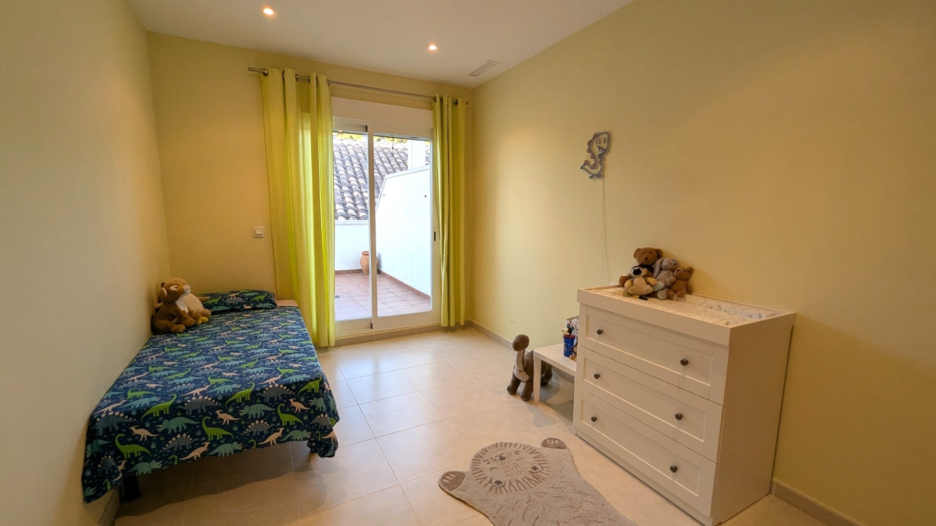 Chambre lumineuse avec sol en grès, rideaux vert citron et porte vitrée coulissante ouvrant sur une terrasse extérieure privée.