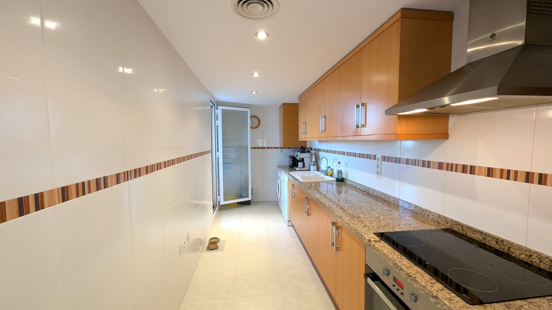 Cuisine indépendante avec meubles en bois, plan de travail en granit et électroménagers intégrés. Murs carrelés blancs avec frise décorative et éclairage par spots encastrés.