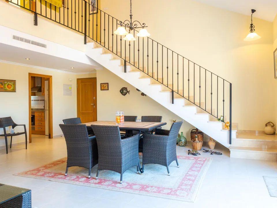Espace salle à manger avec mobilier en osier sombre situé sous un escalier clair à rampe en fer forgé. Pièce de vie lumineuse avec revêtement de sol en carrelage.
