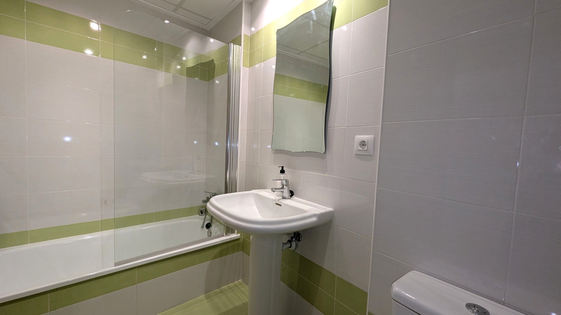 Salle de bain avec baignoire, paroi en verre et lavabo sur colonne, décorée de carreaux blancs et vert citron avec un miroir aux bords ondulés.