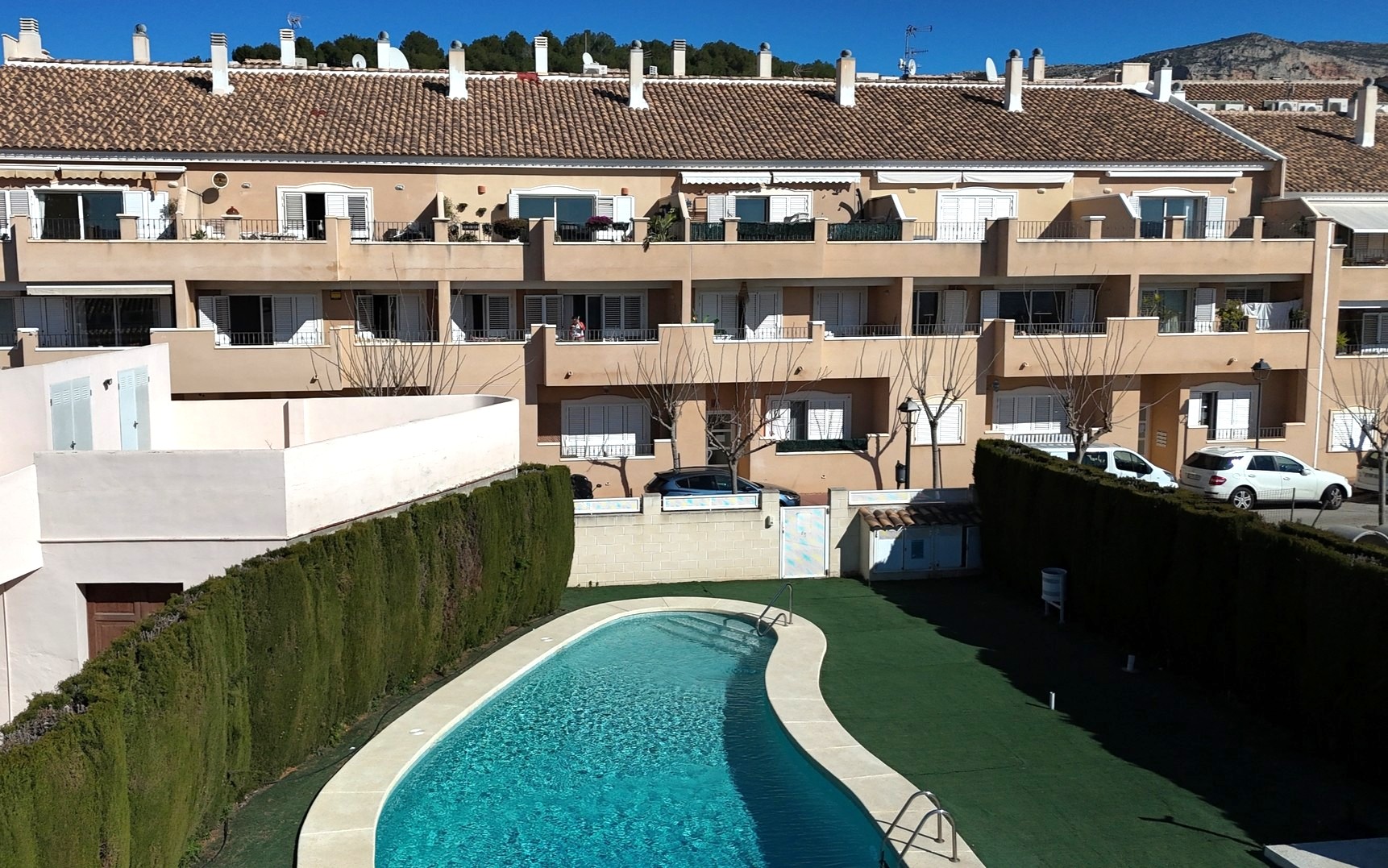 Vue sur la piscine communautaire de forme courbe entourée de gazon synthétique et de hautes haies, avec en arrière-plan la façade aux volets blancs.
