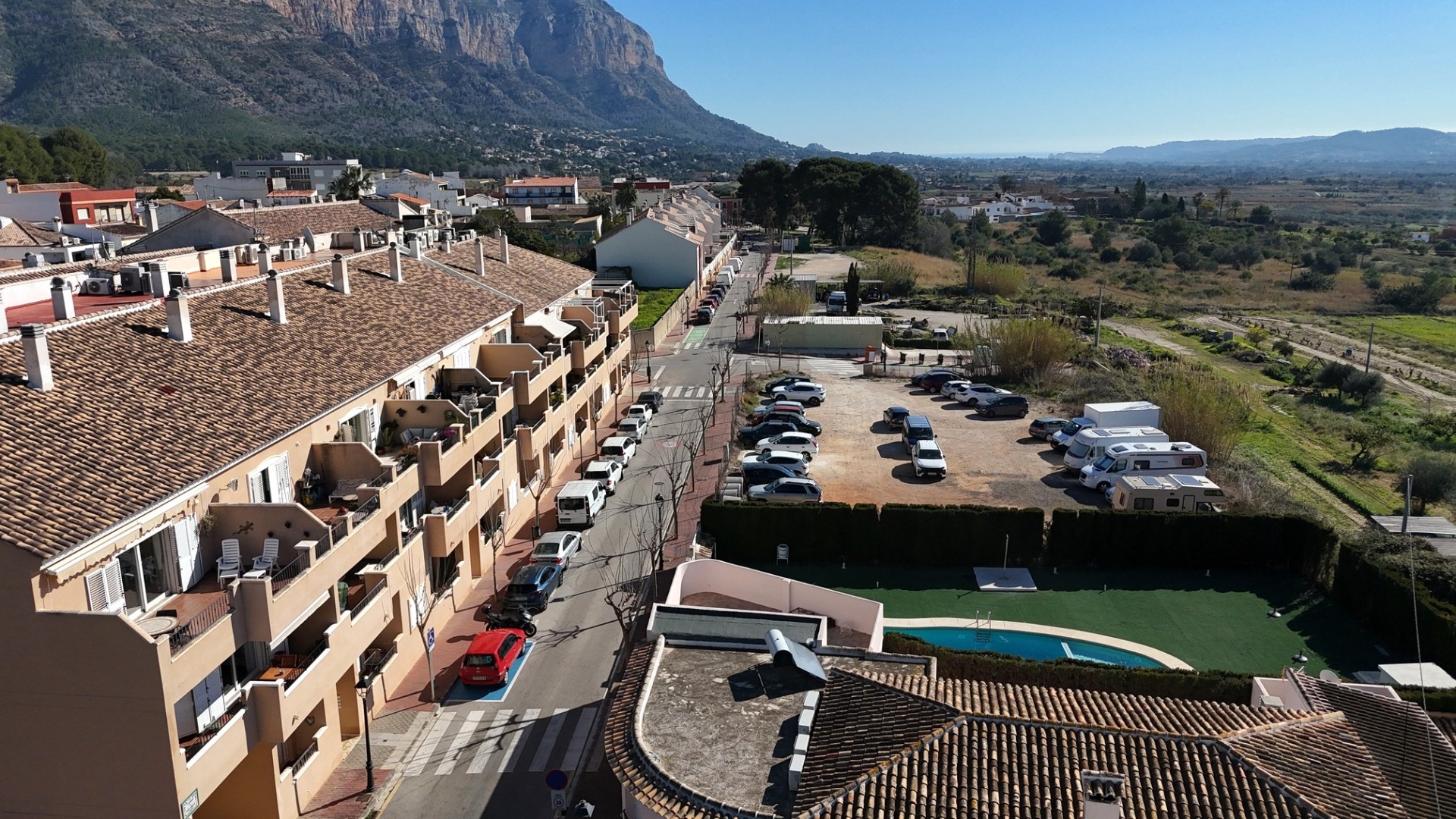 Vue surélevée du complexe résidentiel comprenant la piscine commune, le parking et des vues panoramiques sur le Montgó et la campagne environnante.
