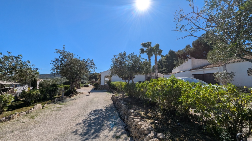 Allée de gravier bordée de murets en pierre traditionnelle menant à la maison principale de style finca, entourée de palmiers et de végétation méditerranéenne.