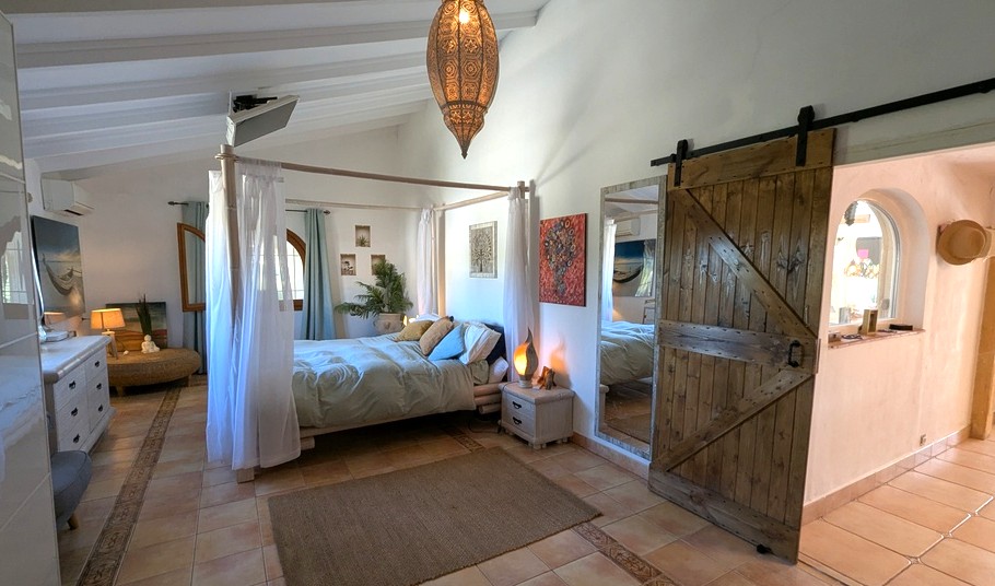 Chambre avec lit à baldaquin, plafonds à poutres blanches et porte coulissante rustique. Le sol en terre cuite et la lampe marocaine soulignent le style méditerranéen.