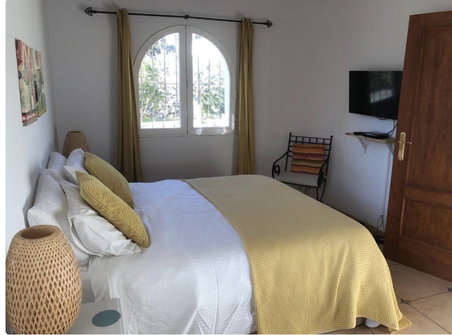 Chambre lumineuse avec fenêtre en plein cintre, sol en terre cuite et télévision murale. La décoration allie des tons moutarde et des lampes de chevet en osier.