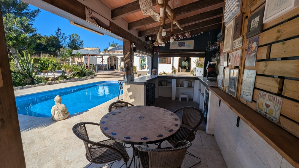 Cuisine d'été couverte avec bar, poutres apparentes et table en mosaïque offrant une vue sur la piscine et les jardins de la propriété.
