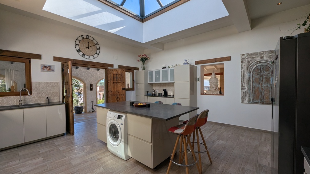 Cuisine moderne avec îlot central et un puits de lumière vitré. L'espace se distingue par ses menuiseries en bois traditionnel et son accès vers la naya arquée.