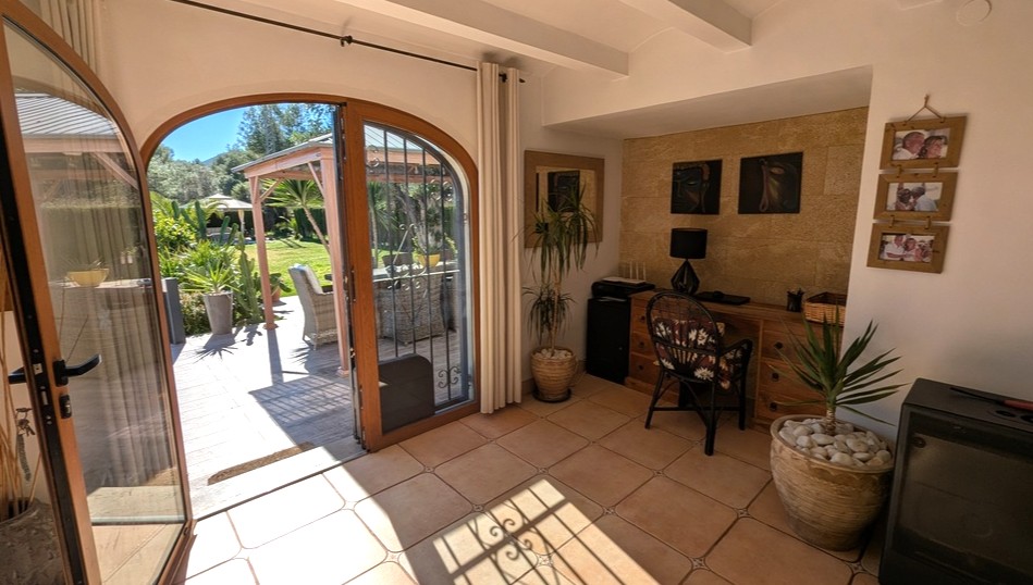Espace bureau lumineux avec sol en terre cuite, mur en pierre naturelle et portes cintrées en bois s'ouvrant sur une terrasse de jardin ensoleillée.