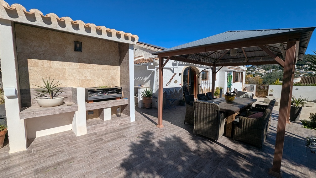 Espace dînatoire extérieur comprenant un barbecue intégré en pierre et une grande pergola abritant une table avec chaises en osier sur un sol en grès cérame.