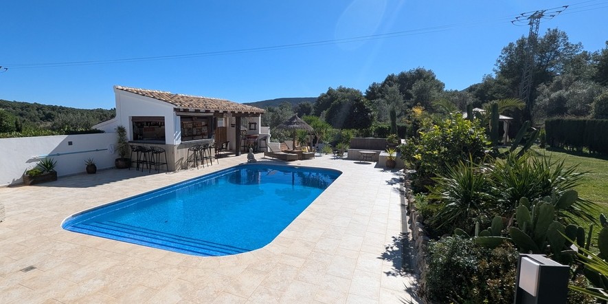 Espace piscine avec terrasse en pierre, cuisine d'été aménagée avec bar, et jardin paysager aux essences méditerranéennes offrant une vue sur la campagne.