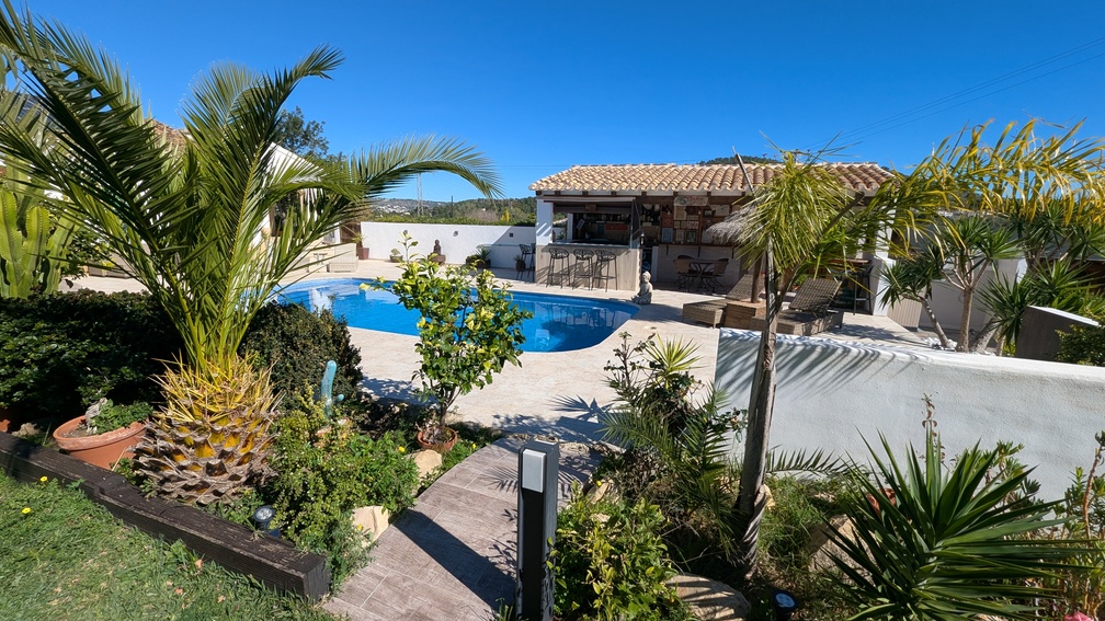 Espace piscine avec terrasse en pierre, jardin méditerranéen planté de palmiers et cuisine d'été couverte équipée d'un bar et de tabourets.