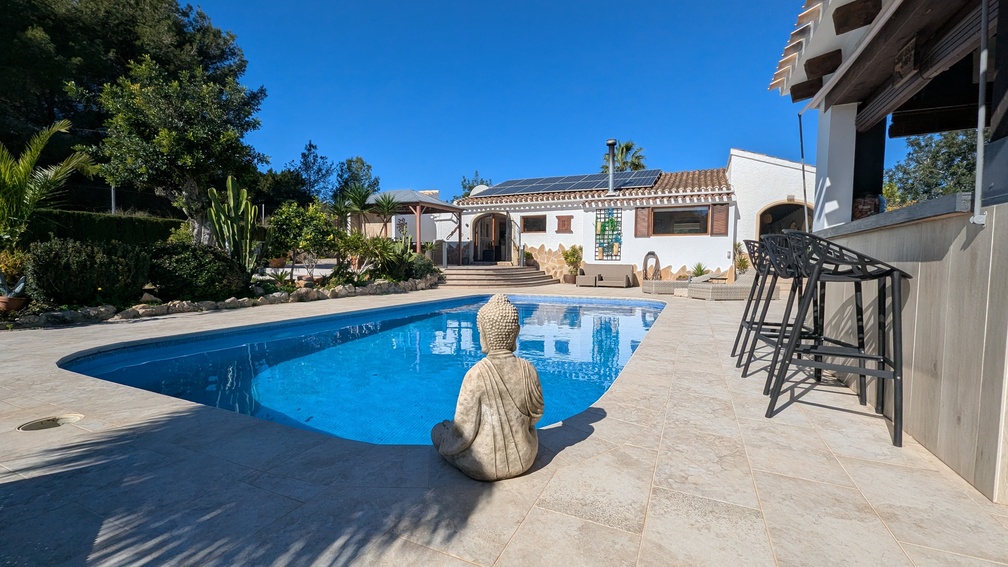 Espace piscine privée avec terrasse en pierre, statue de Bouddha, bar extérieur moderne et villa principale équipée de panneaux solaires.