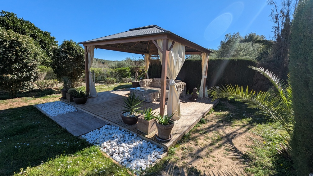 Gazébo en bois sur terrasse avec salon de jardin en rotin, rideaux clairs et bordure de galets blancs au cœur d'un jardin paysager ensoleillé de la Costa Blanca.