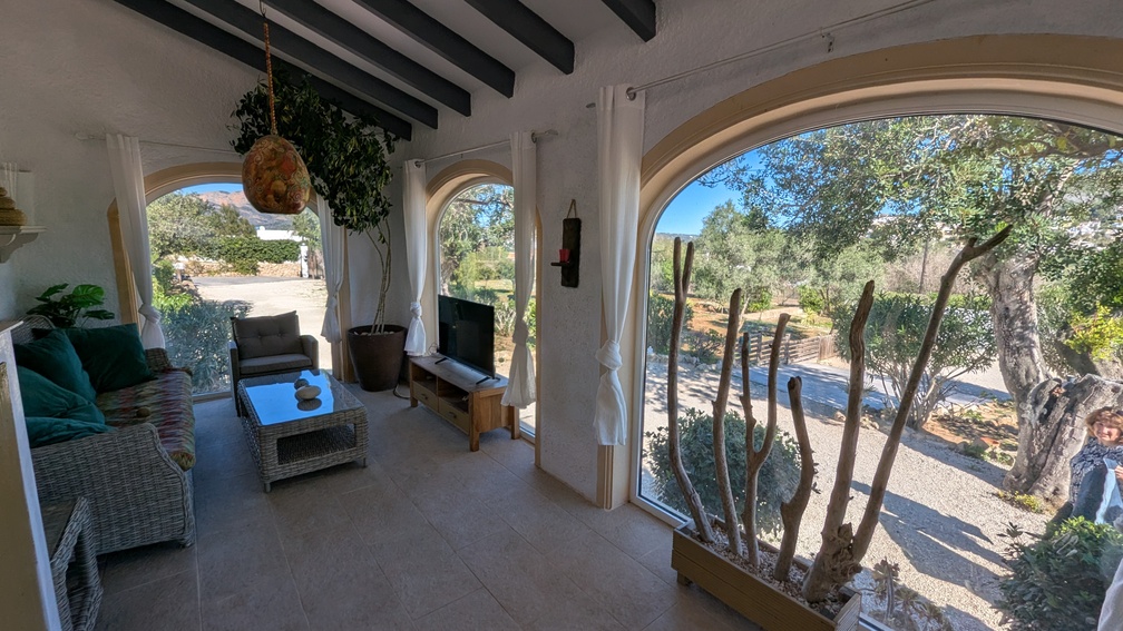 Naya vitrée avec poutres en bois sombre, fenêtres cintrées offrant une vue sur la campagne, et salon avec mobilier en osier et sol en carrelage.