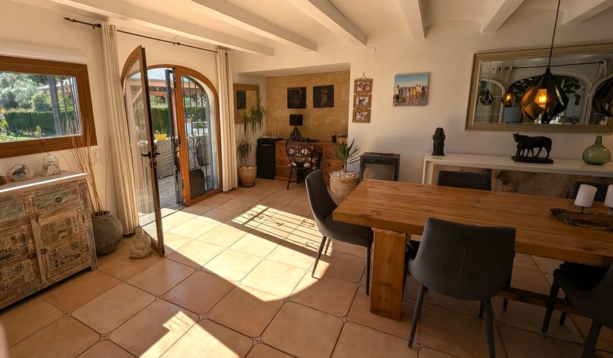 Salle à manger lumineuse avec poutres apparentes blanches, sol en terre cuite et table en bois massif. Une porte cintrée vitrée s'ouvre sur la terrasse et le jardin.