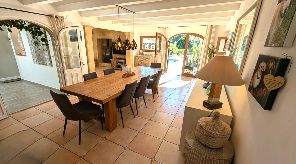 Salle à manger lumineuse avec table en bois massif, poutres blanches et sol en terre cuite. Les portes vitrées en arche s'ouvrent directement sur la terrasse de la piscine.