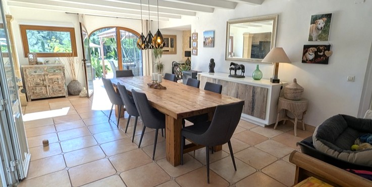 Salle à manger spacieuse avec table en bois massif, poutres apparentes blanches et sol en terre cuite, s'ouvrant sur l'extérieur par une porte cintrée vitrée.
