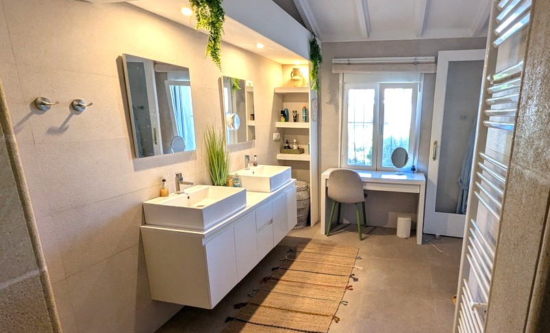 Salle de bain moderne avec double vasque, meuble suspendu et étagères. Comprend un coin coiffeuse sous la fenêtre et un plafond à poutres blanches élégant.