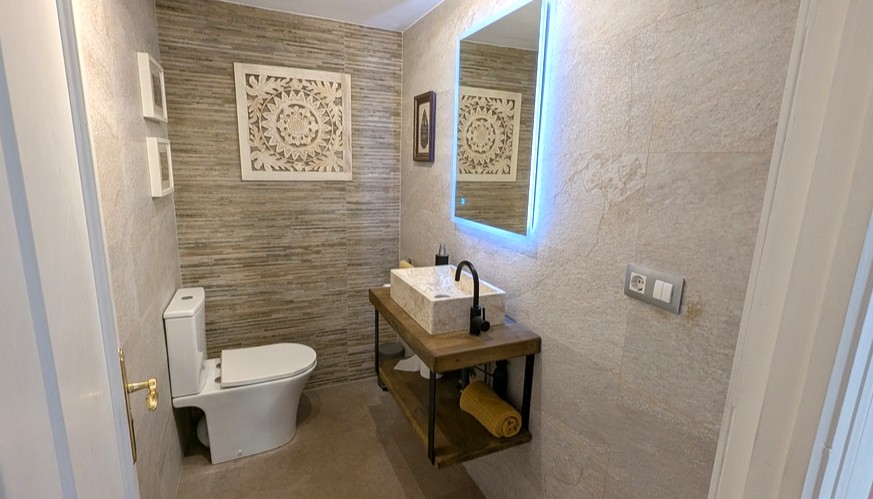 Salle d'eau moderne équipée d'une vasque en pierre sur un meuble suspendu en bois, d'un miroir rétroéclairé et de murs à effet pierre texturée.