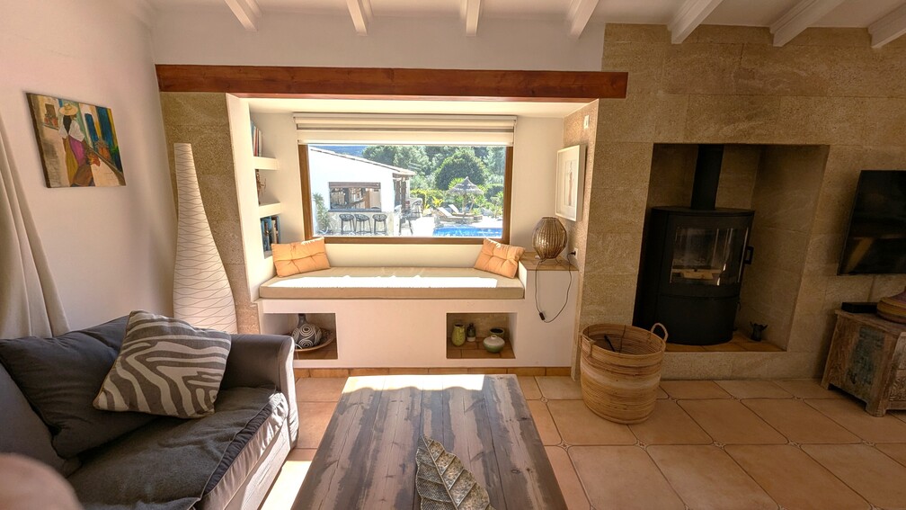 Salon avec banquette intégrée sous la fenêtre offrant une vue sur la piscine, poêle à bois moderne dans un mur en pierre de Tosca et poutres apparentes.