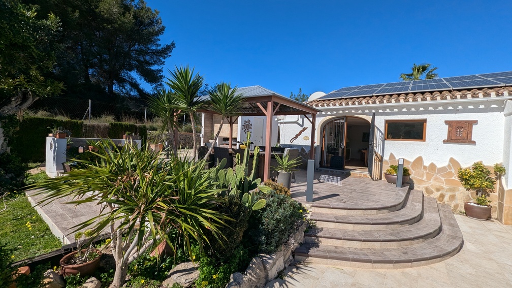 Terrasse avec marches en pierre incurvées, gazebo en bois pour les repas en plein air et façade blanche équipée de panneaux solaires.