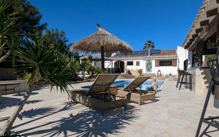 Terrasse carrelée avec piscine privée, transats en osier sous un parasol en chaume et bar d'été. La maison principale est équipée de panneaux solaires en toiture.