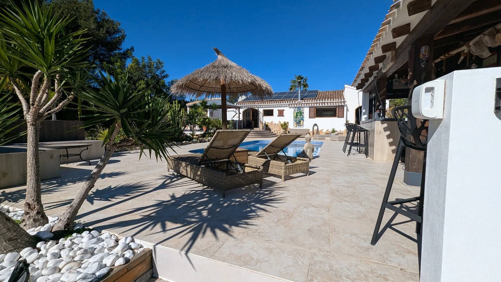 Terrasse ensoleillée avec carrelage en pierre, transats en osier et parasol en paille bordant la piscine, face à la villa principale dotée de panneaux solaires.
