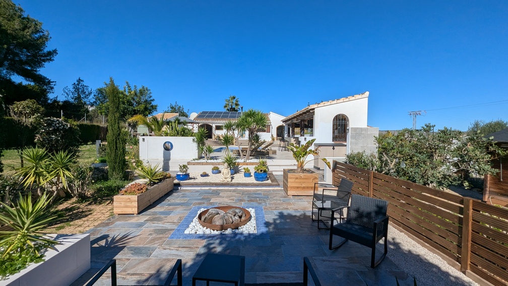 Terrasse extérieure paysagée avec brasero central, dallage en ardoise et escaliers menant à la villa principale équipée de panneaux solaires.
