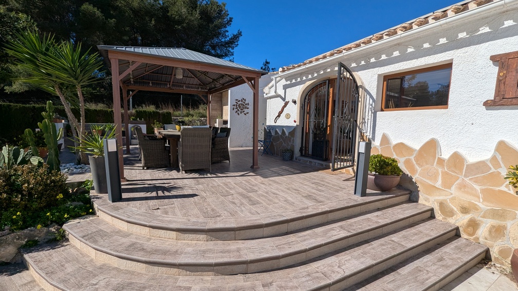 Terrasse surélevée avec carrelage effet pierre et marches courbes menant à un coin repas ombragé sous une pergola en bois, bordée d'une façade blanche aux détails en pierre naturelle.