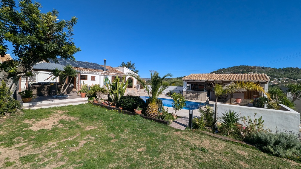 Vue extérieure de la maison principale avec panneaux solaires, piscine privée et cuisine d'été couverte, le tout niché dans un jardin paysager soigné.